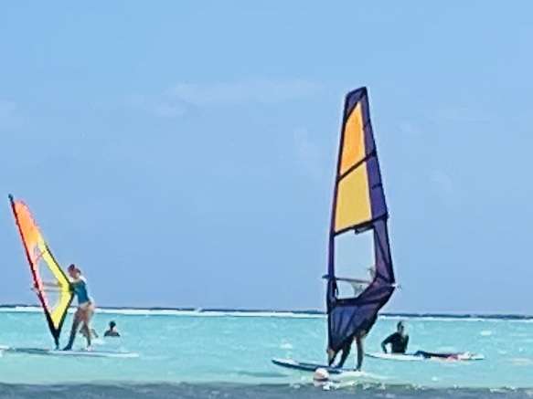Two wind surfers with colorful sails navigatine around several people in the shallow turquoise water